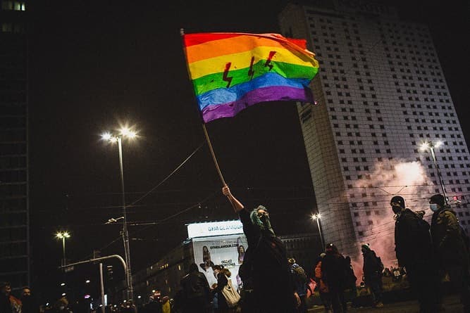 Žmogus protestuojančioje minioje laiko iškeltą LGBT+ vėliavą su trimis raudonais žaibais.
