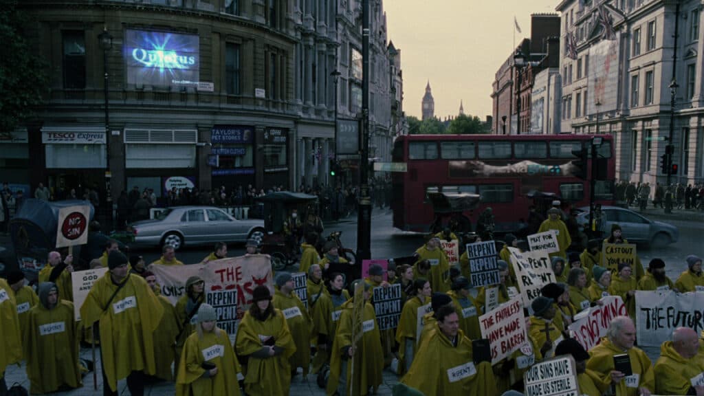 Pilkas distopinis Londonas, kuriame protestuoja geltonais lietpalčiais apsirengę žmonės