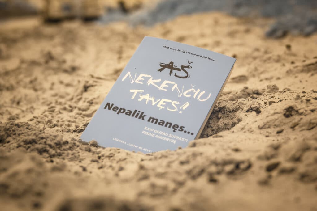 smėlyje gulinti knyga "Aš nekenčiu tavęs! Nepalik manęs"