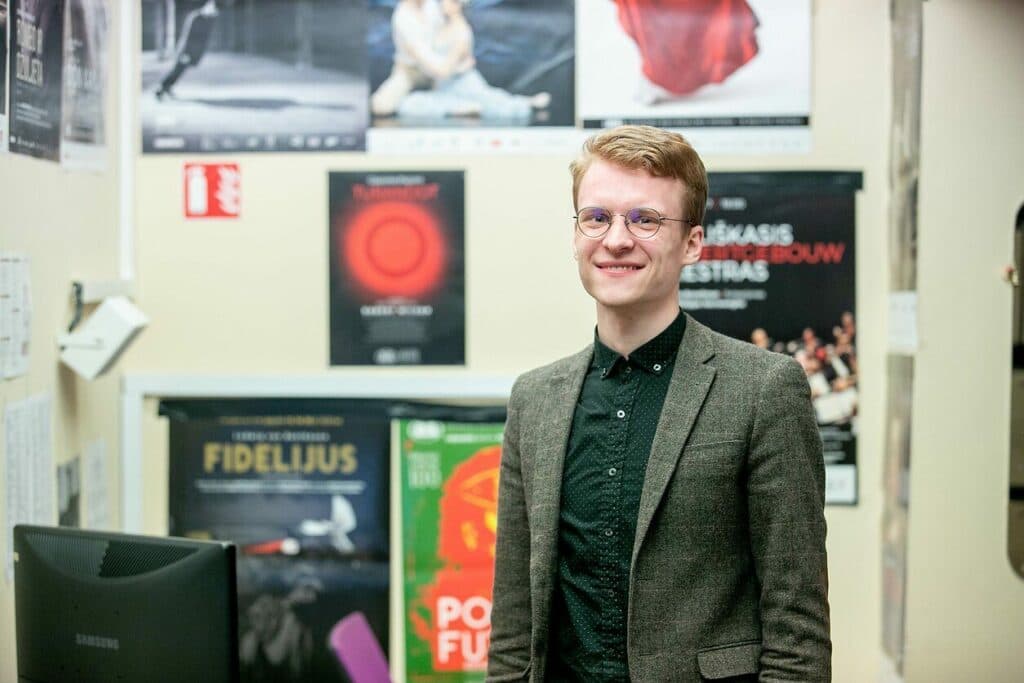 Teatro bibliotekininkas stovi prie sienos su spektaklių plakatais